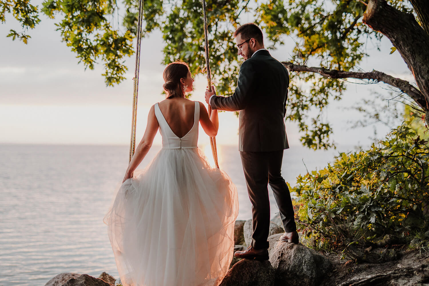 Para młoda uchwycona na naturalnej fotografii. Panna młoda siedzi na huśtawce, obok stoi pan młody. Patrzą sobie w oczy.