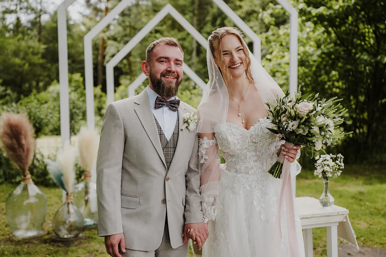 Przykładowy kadr przedstawiający ofertę fotografii ślubnej, na którym para młoda pozuje uśmiechnięta w miejscu ceremonii ślubnej.